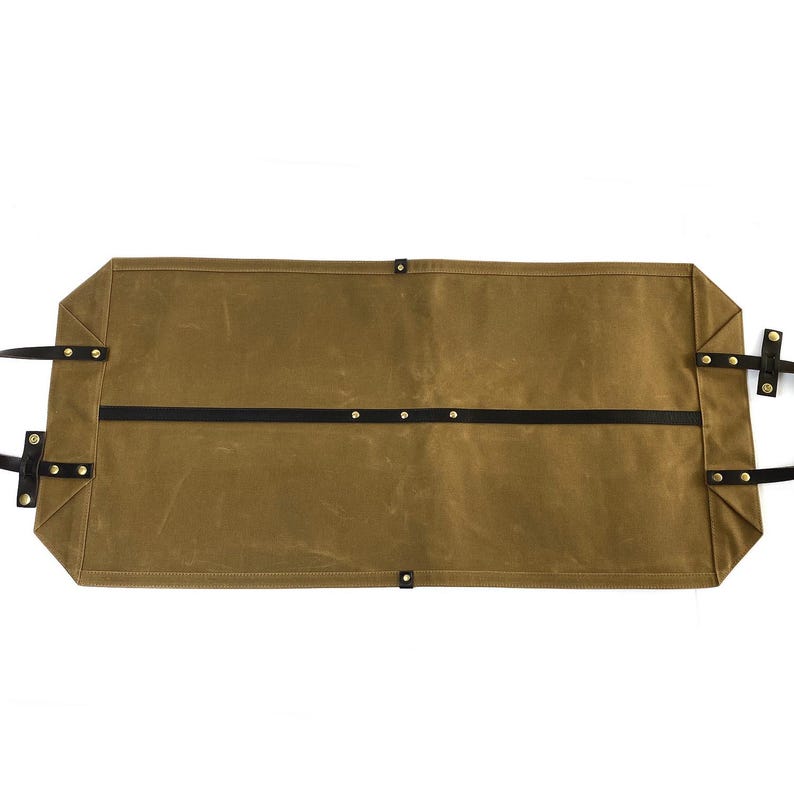 May include: A brown waxed canvas tool roll with black leather straps and brass rivets. The roll is rectangular with a flat bottom and top, and has two black leather straps running across the middle. The handles are black leather.