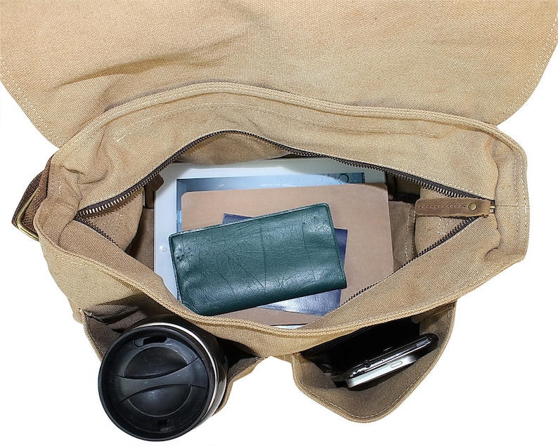 May include: A tan canvas messenger bag with a zippered top and a brown leather flap. The bag is open and contains a tablet, a notebook, a wallet, a phone, and a travel mug.