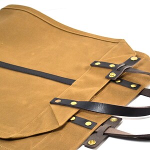 May include: A tan canvas tote bag with black leather straps and gold-colored rivets. The bag has a rectangular shape with a curved top edge and two leather handles. The bag is designed for carrying items.
