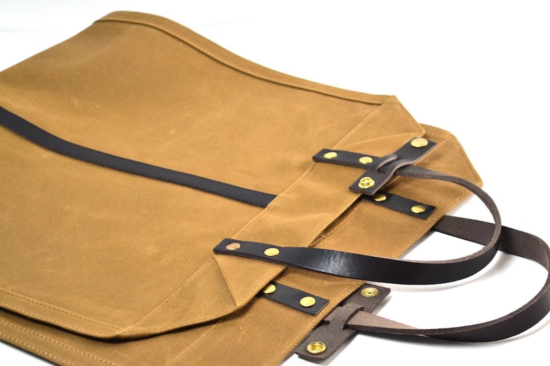 May include: A tan canvas tote bag with black leather straps and gold-colored rivets. The bag has a rectangular shape with a curved top edge and two leather handles. The bag is designed for carrying items.