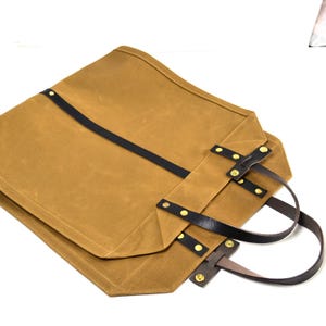 May include: A tan canvas tote bag with black leather straps and handles. The bag features brass rivets and a rectangular shape. The handles are made of brown leather. The bag is folded over.