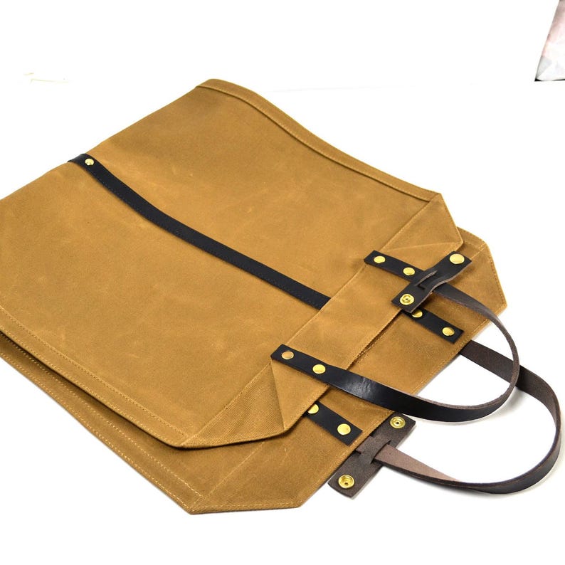 May include: A tan canvas tote bag with black leather straps and handles. The bag features brass rivets and a rectangular shape. The handles are made of brown leather. The bag is folded over.