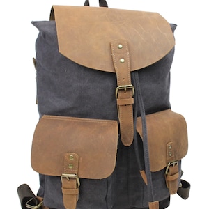 May include: A gray canvas backpack with brown leather accents and buckles. The backpack has two large front pockets with buckles and a drawstring closure at the top.