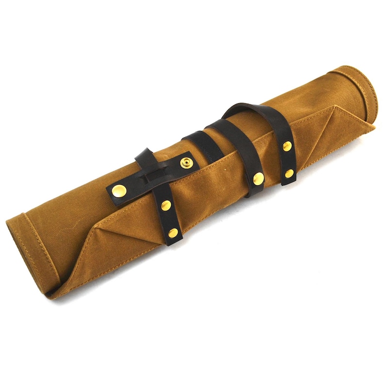 May include: A rolled-up brown canvas tool roll with black leather straps and gold-colored rivets. The tool roll is secured with two leather straps and a buckle. The canvas has a textured appearance.