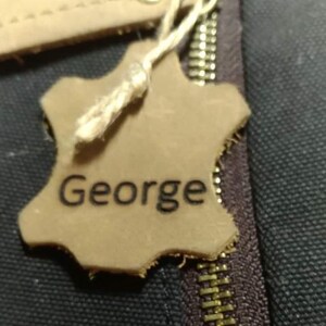 May include: A close-up of a leather tag with the name "George" printed on it. The tag is attached to a zipper with a gold-colored pull. The tag is a light brown color and has a rough, uneven edge.