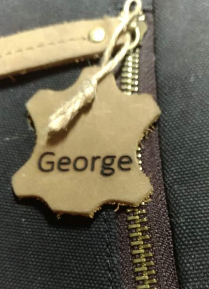 May include: A close-up of a leather tag with the name "George" printed on it. The tag is attached to a zipper with a gold-colored pull. The tag is a light brown color and has a rough, uneven edge.