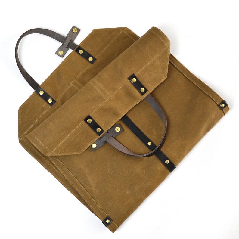 May include: A brown waxed canvas tote bag with black leather straps and brass rivets. The bag has a rectangular shape with a folded flap and a handle for carrying. The bag is designed for carrying items.