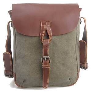 May include: A canvas messenger bag with a brown leather flap and accents. The bag features adjustable brown leather straps and a buckle closure. The main body of the bag is olive green.
