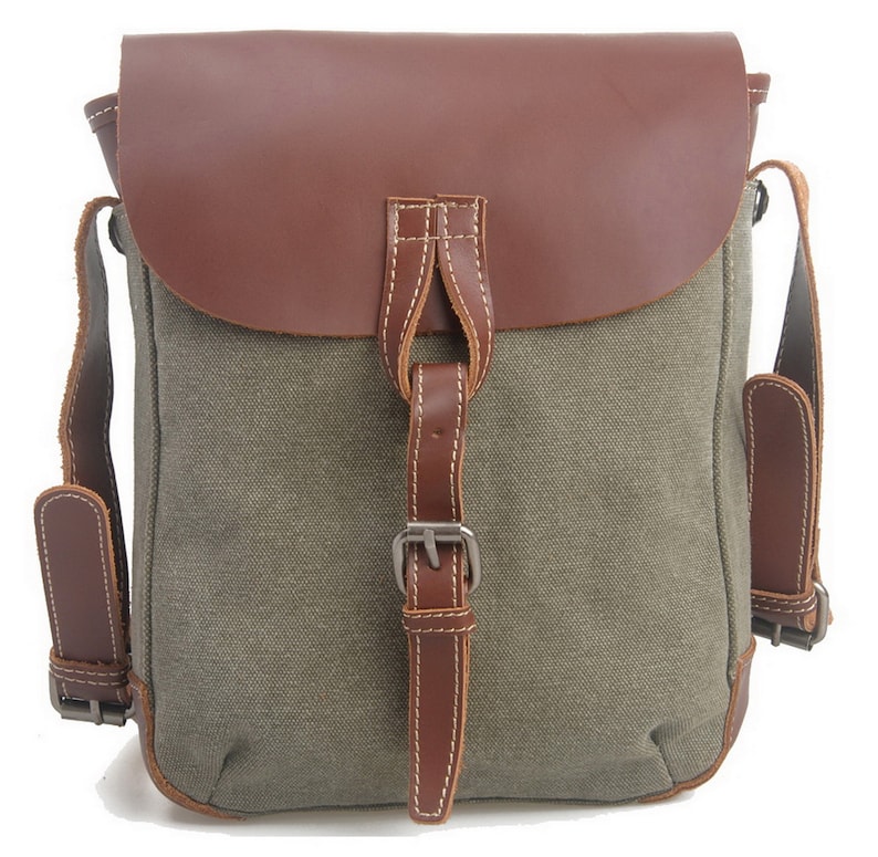 May include: A canvas messenger bag with a brown leather flap and accents. The bag features adjustable brown leather straps and a buckle closure. The main body of the bag is olive green.