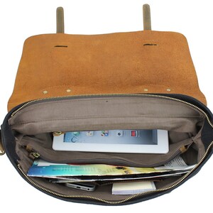 May include: Open black canvas and brown leather messenger bag with a zipper closure. The bag is lined with a gray fabric and has a zippered pocket on the inside. The bag is filled with a tablet, magazines, and other items.