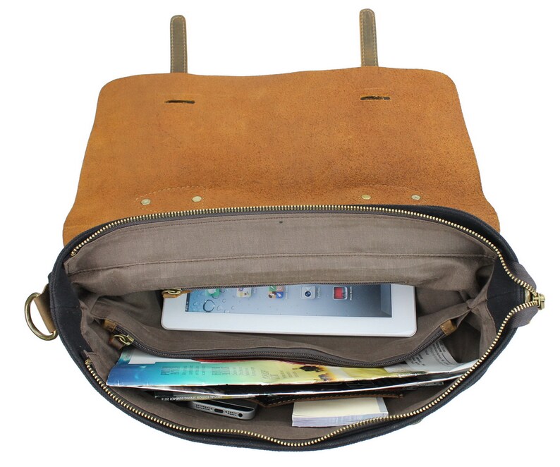 May include: Open black canvas and brown leather messenger bag with a zipper closure. The bag is lined with a gray fabric and has a zippered pocket on the inside. The bag is filled with a tablet, magazines, and other items.