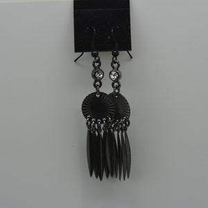 May include: Black dangle earrings with a round, textured disc and a crystal accent. The earrings have a fringe of small, pointed metal pieces.