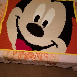 May include: A crocheted blanket featuring a cartoon character with a red nose, black eyes, and a big smile. The blanket is framed with a yellow border and has a red and orange background.