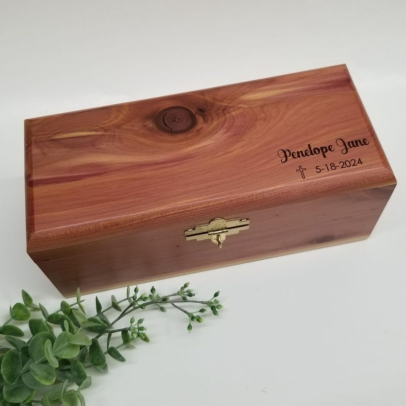 May include: A wooden box with a brass latch. The box is engraved with the name "Penelope Jane" and the date "5-18-2024". A small cross is engraved above the date.