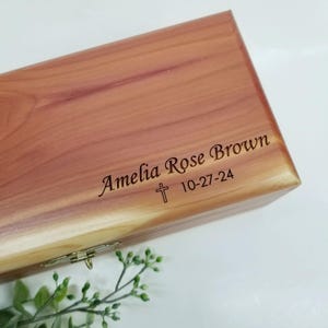 May include: A wooden box with a hinged lid, engraved with the name "Amelia Rose Brown" and the date "10-27-24". A small cross is engraved above the date.