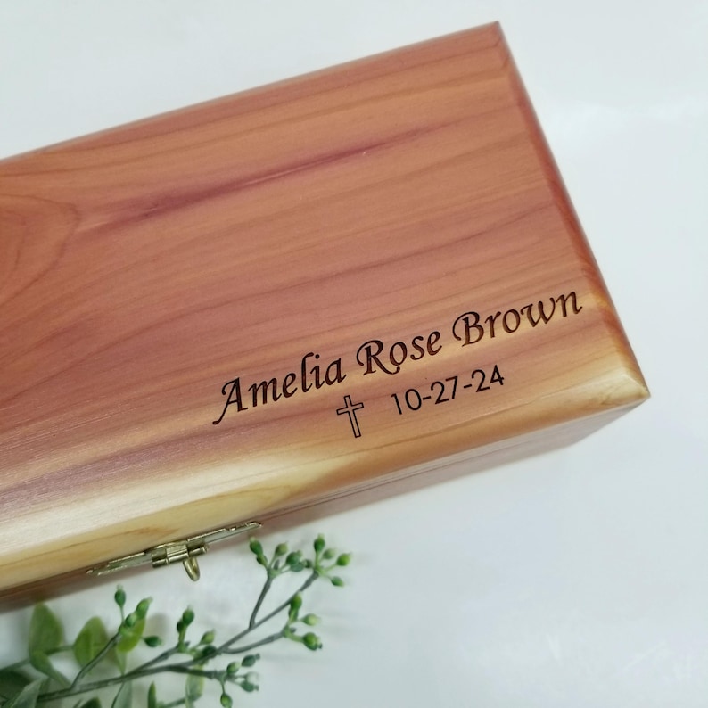 May include: A wooden box with a hinged lid, engraved with the name "Amelia Rose Brown" and the date "10-27-24". A small cross is engraved above the date.