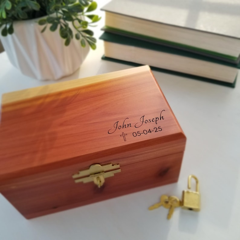 May include: A wooden box with a brass latch and a keyhole. The box is engraved with the name "John Joseph" and the date "05-04-25". A small cross is engraved above the date.