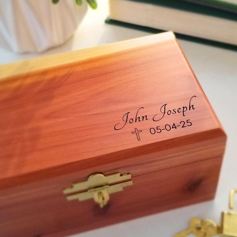 May include: A wooden box with a hinged lid and a brass latch. The box is engraved with the text "John Joseph 05-04-25" and a small cross.