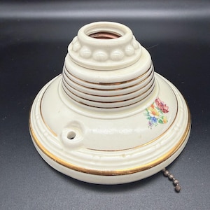 May include: Cream-colored vintage ceiling light fixture with gold trim and a floral design. The fixture has a decorative base with a pull chain and a bulb socket. The fixture is made of ceramic and has a classic, antique look.