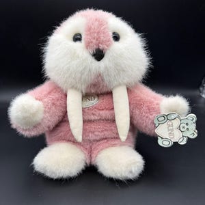 May include: A plush walrus toy featuring soft pink and white fur, two long white tusks, and dark eyes. A silver-toned oval tag with the text GUND is attached to the chest, and a light blue bear-shaped tag with the text GUND is attached to the arm.