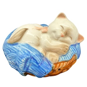Puede incluir: Figura de cerámica de un gato durmiendo. El gato es blanco con orejas y patas marrones y un lazo rosa. Descansa sobre un cojín azul dentro de una cesta naranja. La figura tiene un acabado brillante.