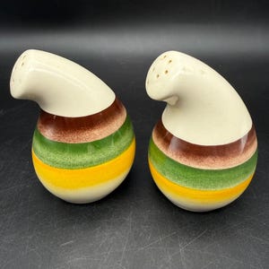May include: A pair of ceramic salt and pepper shakers with a curved, teardrop-like shape. Each shaker features a cream-colored top with small holes and a body decorated with horizontal bands of brown, green, and yellow glaze over a cream base.