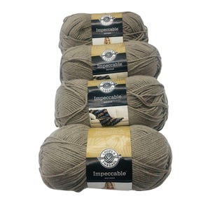 May include: Four skeins of Loops & Threads Impeccable worsted yarn in a neutral taupe color. Each skein is labeled and stacked vertically, showcasing the yarn's texture and brand.