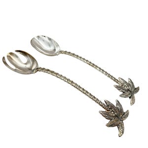 Puede incluir: Un par de cubiertos de ensalada plateados. Uno es una cuchara, el otro un tenedor. Ambos tienen mangos texturizados y diseños de hojas de palmera en el extremo. Los cubiertos están sobre un fondo blanco.