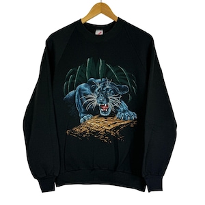 Op de afbeelding: Zwart sweatshirt met een gedetailleerde illustratie van een panter met blauwe accenten, die zijn tanden laat zien. De panter staat tegen een achtergrond van groen gebladerte en een bruine boomstam. De sweatshirt heeft een ronde hals en lange mouwen.