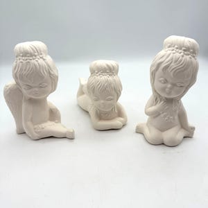 Op de afbeelding: Drie ongeverfde keramische cherubijnbeeldjes in verschillende poses. Eén cherubijn zit met vleugels, een andere ligt en de derde zit met een hand op de borst. Elk heeft een kroonachtig hoofddeksel en gedetailleerde gelaatstrekken.