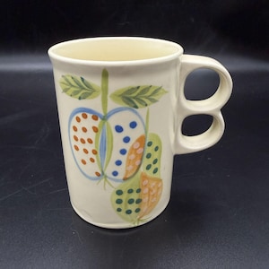 May include: Cream-colored ceramic mug with a whimsical fruit design. The mug features a handle with two finger holes. The fruit illustrations include apples and pears in shades of blue, orange, and green, with green leaves. The mug has a slightly flared shape.