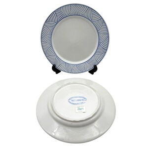 May include: Two white ceramic plates. One plate is upright on a black stand, displaying a blue wave pattern around the rim. The other plate is face down, revealing a stamp with text. A classic design for dining.