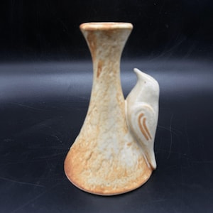 Può includere: Un vaso in ceramica con un design a forma di uccello. Il vaso è beige con dettagli marroni e una superficie strutturata. L'uccello è appollaiato sul lato del vaso, con dettagli alari marroni. Il vaso ha una base svasata e un collo stretto.