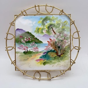 May include: A square, hand-painted ceramic tile featuring a landscape scene with trees, water, and mountains. The artwork is framed by a decorative, gold-colored metal wire frame. The painting includes a signature in the bottom right corner.