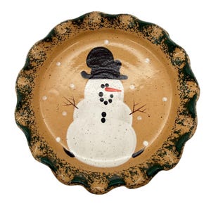May include: A ceramic pie plate with a snowman design. The plate has a tan background with a green scalloped edge. The snowman has a black top hat, coal eyes, and a carrot nose. Snowflakes are scattered around the snowman.