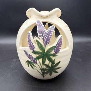 Puede incluir: Un jarrón de cerámica de color crema con un diseño redondeado y calado que presenta un borde superior festoneado. La parte frontal está decorada con una ilustración pintada a mano de flores de lupino moradas con hojas verdes sobre un fondo blanco roto.