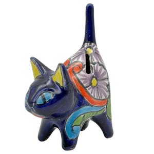 May include: A ceramic cat-shaped piggy bank in a vibrant blue hue. The cat features yellow-tipped ears, blue eyes, and a tall tail. The body is adorned with floral and swirl patterns in red, purple, and green. A slot is visible on the back.