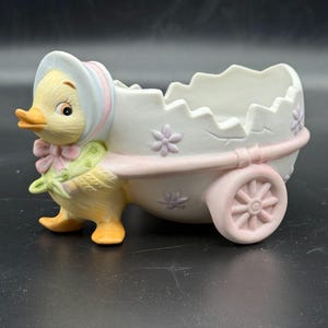 May include: A ceramic figurine of a yellow duckling wearing a bonnet and a green scarf, pulling a white egg-shaped cart with a jagged top edge. The cart features small purple flower accents and pink wheels, with a matte finish throughout the piece.