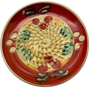 May include: A ceramic grater plate with a red rim and a cream-colored center. The center features a textured grating surface with green, red, and yellow accents. Decorative elements include red and green floral-like shapes and cream-colored dots around the edge.