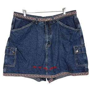 May include: Blue denim skirt with a decorative embroidered waistband and hem. The skirt features two cargo pockets and red embroidered accents near the hem. The skirt is hanging on a metal hanger.
