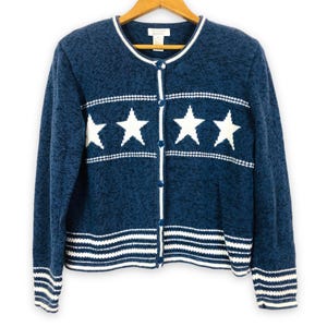 Könnte beinhalten: Eine dunkelblaue Strickjacke mit Rundhalsausschnitt, langen Ärmeln und einer Knopfleiste. Das Kleidungsstück zeigt ein horizontales Band aus vier weißen Sternen auf der Brust sowie weiße Streifenmuster an den Ärmelbündchen und am Saum.