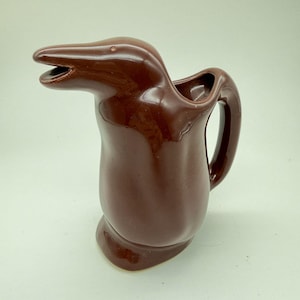 May include: A brown ceramic pitcher with a dolphin-shaped spout and handle. The pitcher has a glossy finish and a unique design, perfect for serving beverages. The dolphin's mouth is open, adding a playful touch to the piece.