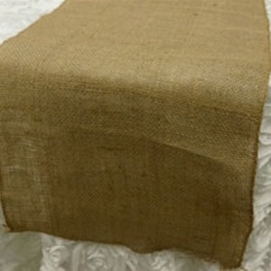 May include: A natural burlap table runner. The runner is a light brown color and is draped over a white, textured surface. The burlap has a coarse weave and a rustic appearance, suitable for event decor or home use.