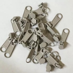 Puede incluir: Un montón de tiradores de cremallera de plástico gris. Los tiradores son todos del mismo tamaño y forma, y tienen un pequeño bucle de metal en la parte superior. Los tiradores están dispuestos en un orden aleatorio.