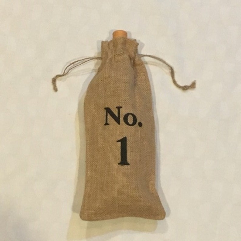 以下が含まれることがあります： 木製のトップと前面に「No. 1」の文字が印刷された、茶色のジュート製のワインボトルバッグです。