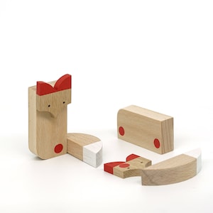 May include: Wooden toy fox puzzle with five pieces. The fox is made of natural wood with red accents and white painted details. The pieces fit together to form a complete fox.