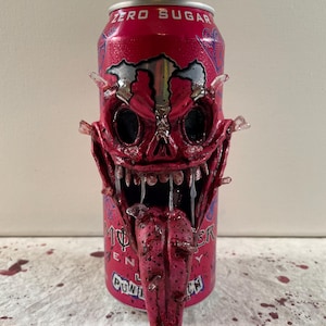 May include: A red Monster Energy drink can with a sculpted, demonic face attached. The face has large, black eye sockets, sharp teeth, and a long, textured tongue. The can reads "ZERO SUGAR" and "MONSTER ENERGY".