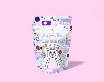 Könnte beinhalten: Ein lila-weißer Beutel mit einem Comic-Kaninchen, das ein Osterei und einen Vogel hält. Der Beutel trägt die Aufschrift "Poppymint Pals Foaming Bunny Bombs" und den Text "Bunny Delights, Natural Bubbles, No Parabens".