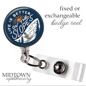 Op de afbeelding: Een marineblauwe badge reel met de tekst "LIFE IS BETTER ON THE SLOPES" en een skiër-afbeelding. Het heeft een witte plastic band met een metalen druksluiting. De tekst "fixed or exchangeable badge reel" is ook zichtbaar.
