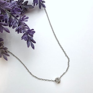 Puede incluir: Un delicado collar plateado con un pequeño colgante hexagonal con la letra "S". La fina cadena y el colgante están centrados. Ramas de lavanda en la esquina superior izquierda, sobre un fondo blanco.